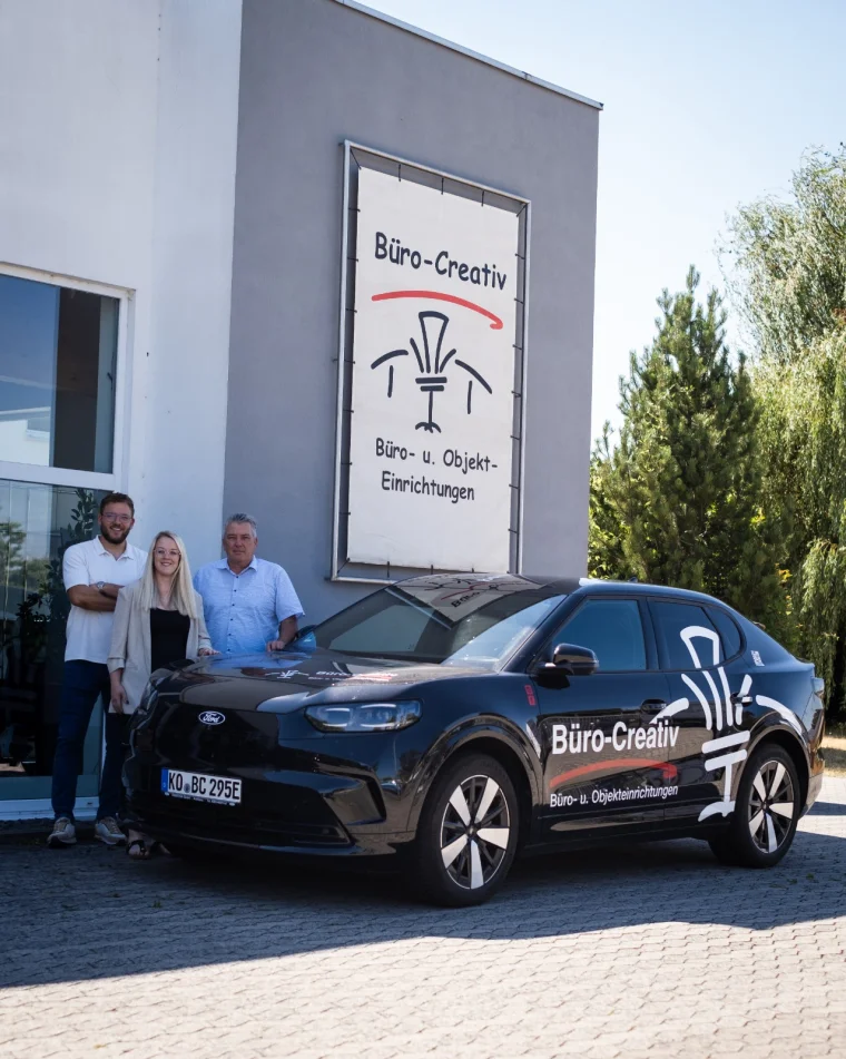 Drei Mitarbeiter von Büro-Creativ vor dem Firmengebäude in Koblenz, daneben der beklebte Firmenwagen unter dem Banner „Büro- u. Objekt-Einrichtungen“